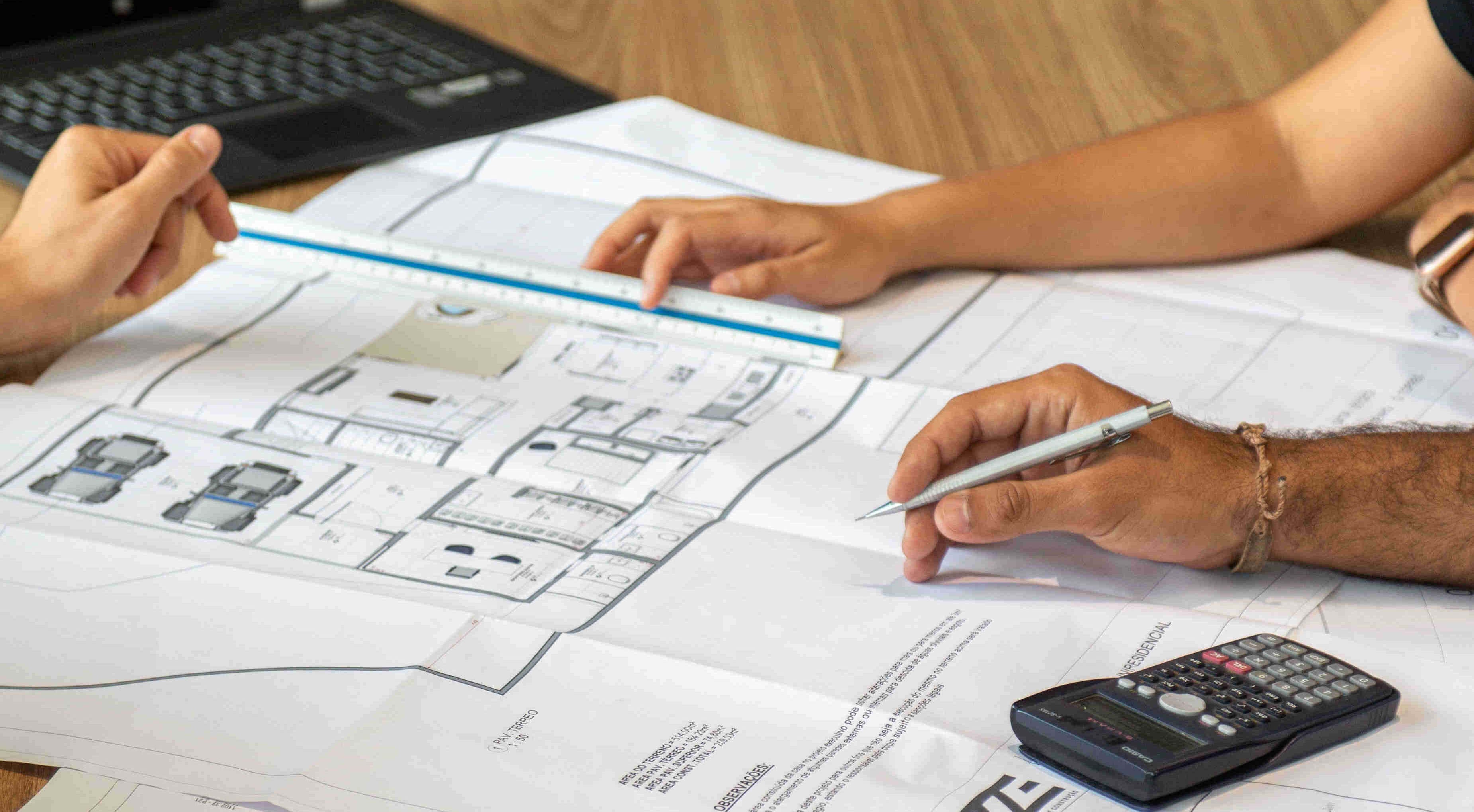 The hands of several people rest on a floor plan; one holds a ruler while another notes figures from a calculator onto the plan with a ballpoint pen.