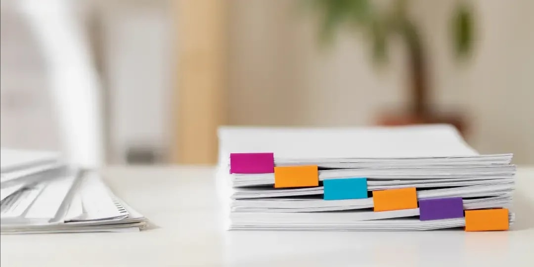 A stack of documents on a white table, with the individual documents held together with colorful clips, and another stack of documents lying next to it