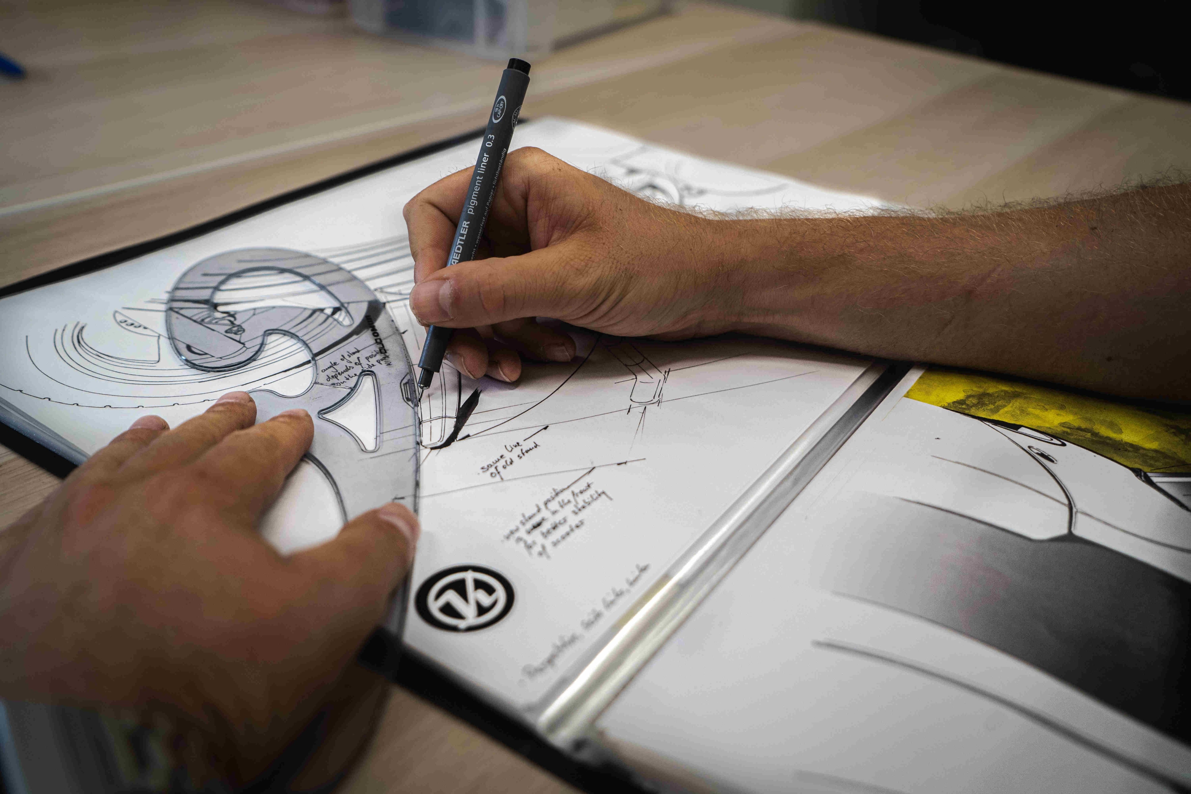 Close-up of hands drawing curved lines into a floor plan using a French curve ruler, part of a portfolio folder; an architect's logo is visible in the corner.