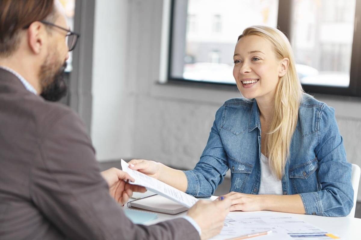 An insurance broker sits across from a female client at a desk and goes through documents with her.