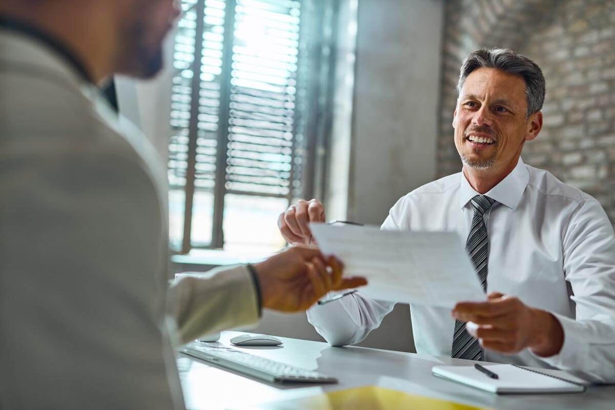 An insurance broker sits across from a client at a desk, smilingly accepting documents from them.