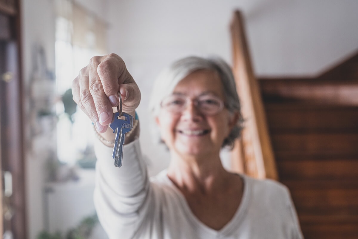 An elderly woman stands smiling in a house entrance, holding up a front door key.