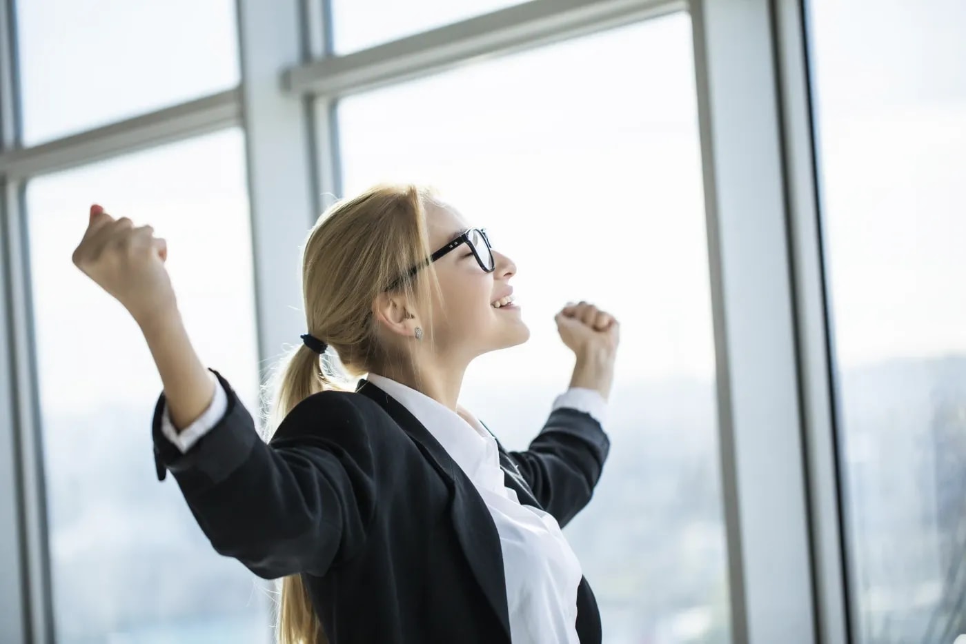 A blonde woman wearing glasses strikes a triumphant victory pose in front of a window front, arms and fists raised, with a very satisfied laugh