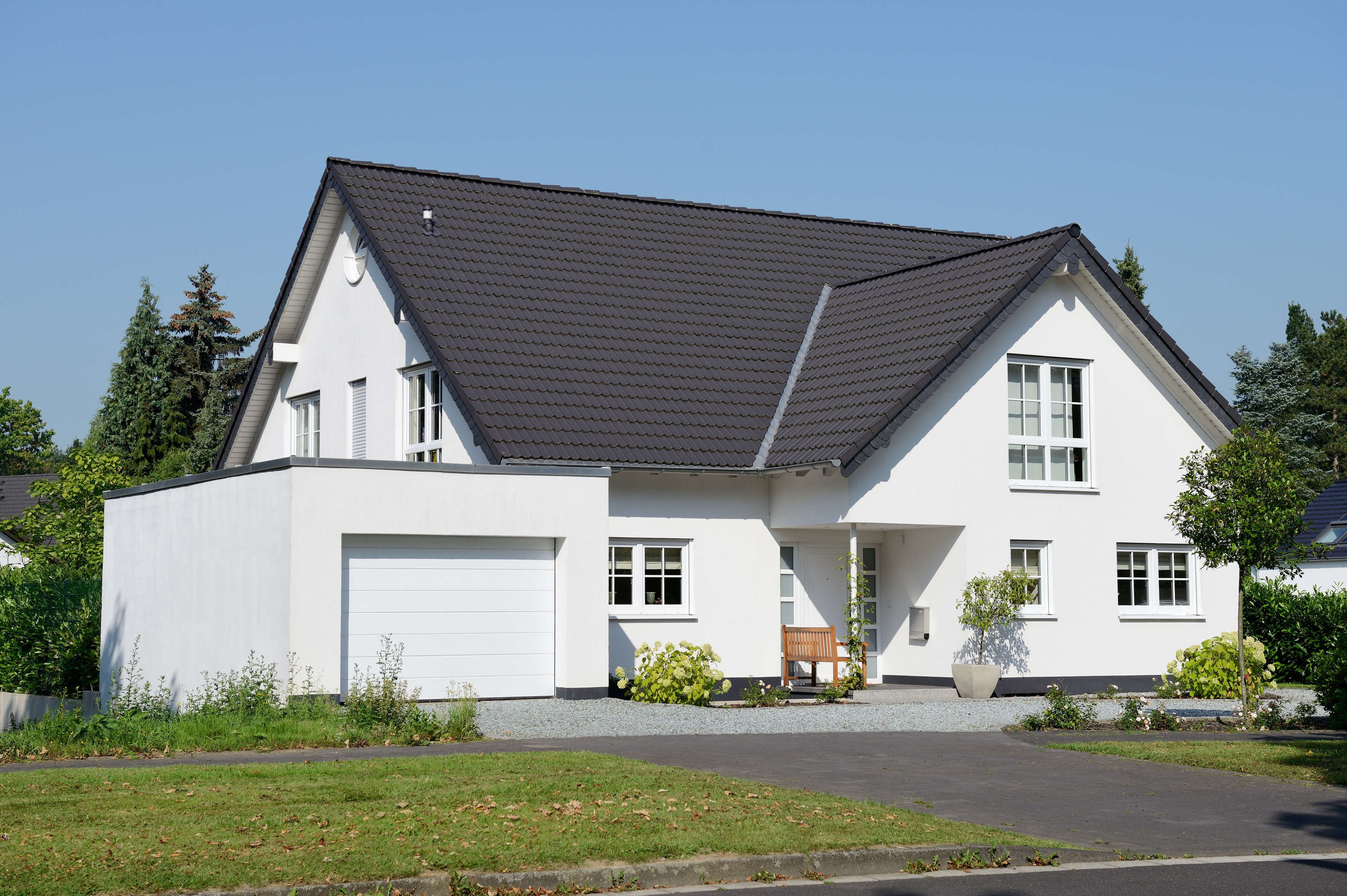 Multi-storey single-family house with an L-shaped footprint, pitched roof and flat-roof garage in the foreground, set between green areas.