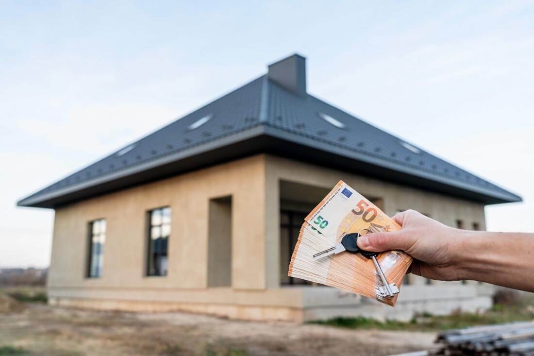 Hand holding euro banknotes and a house key in front of a newly built bungalow