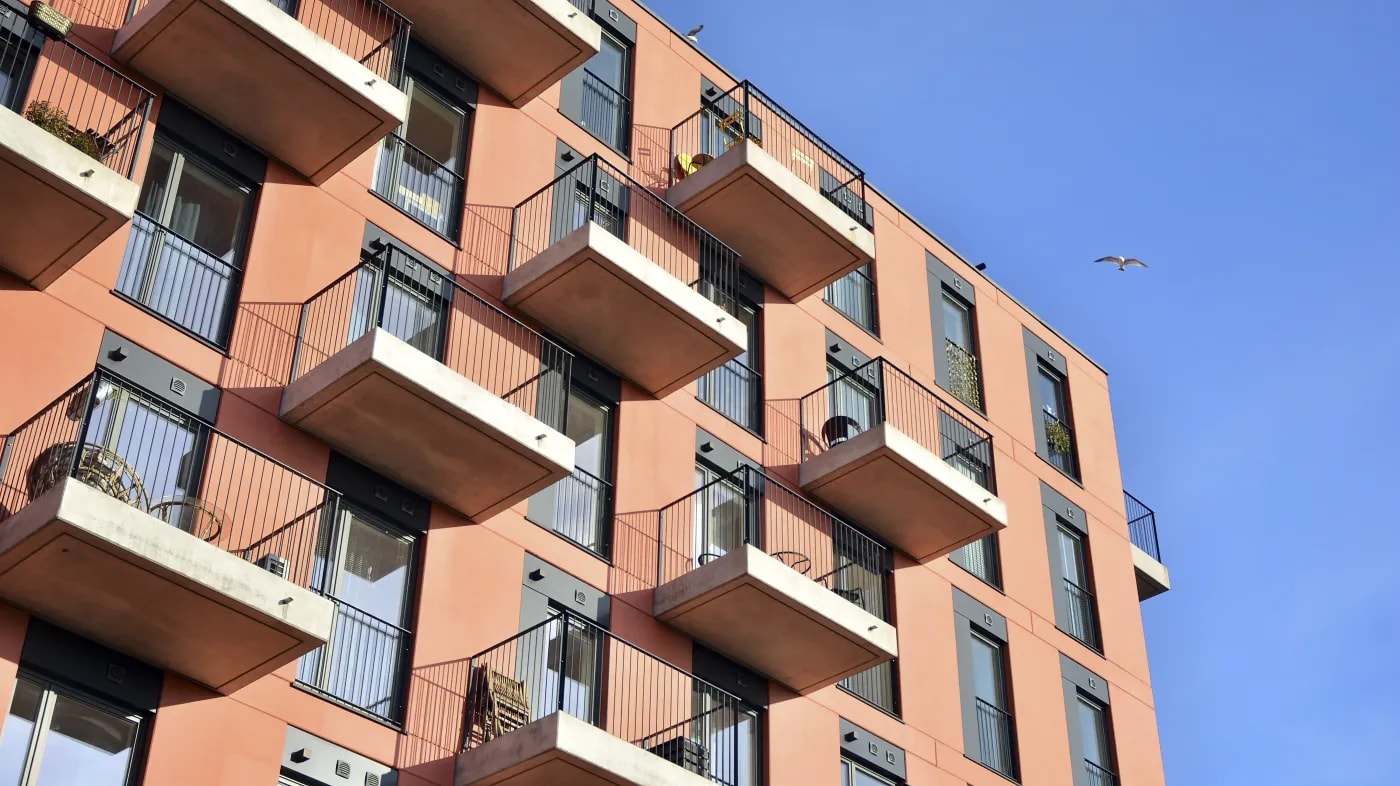 Ansicht von unten auf die rechte obere Ecke der Fassade eines mehrgeschossigen Stadt-Mehrfamilienhauses mit um je eine Fensterbreite versetzten Balkonen je Wohneinheit.
