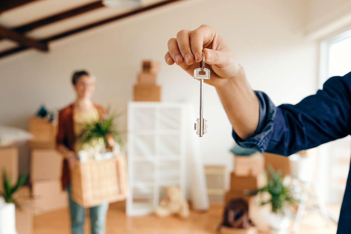 Hand holding a front door key towards the camera, with a woman carrying a moving box in the background during a house move