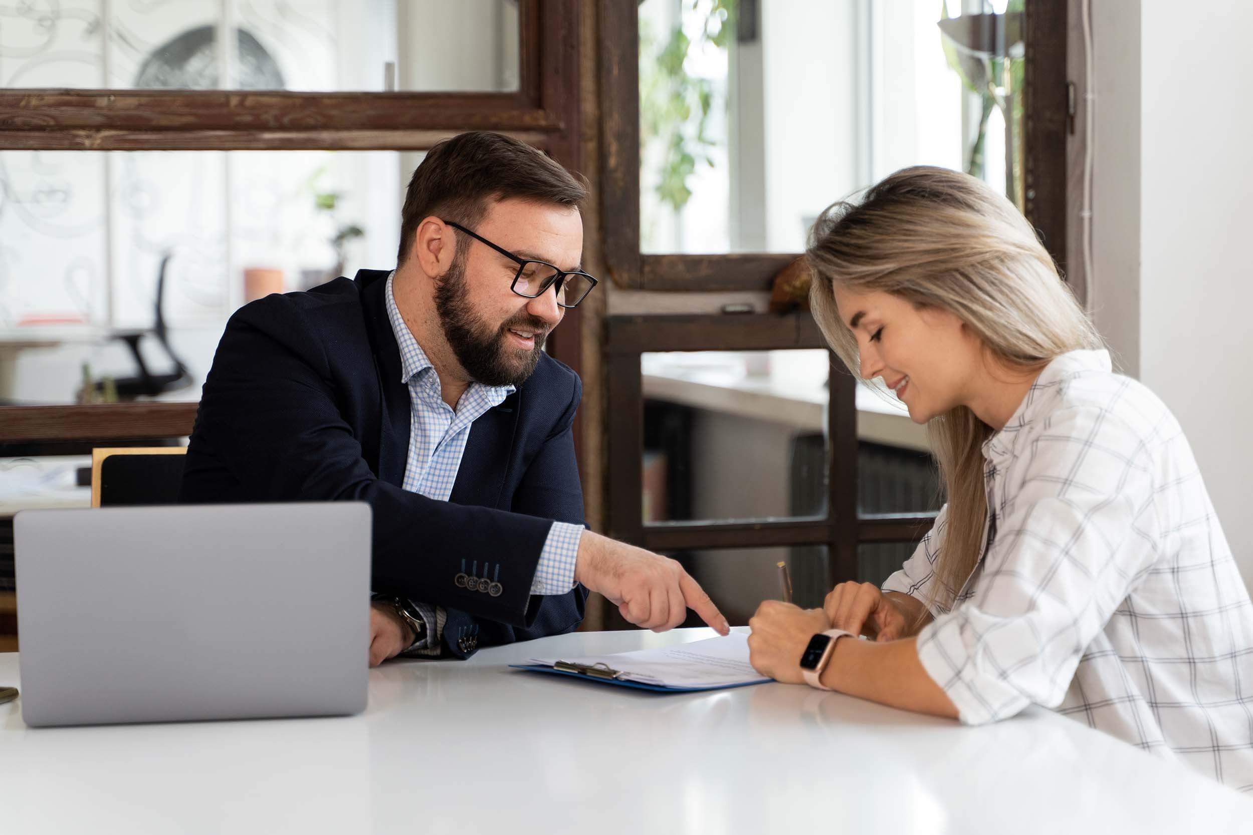 A tax advisor with a laptop sits with a client at a meeting table, explaining a form on a clipboard to her