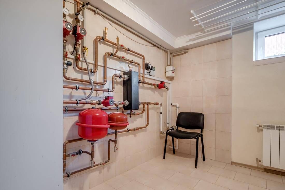 View of a basement room from the entrance, with some heating pipes along the left wall and a chair in the back corner beneath a half-window at the top of the adjacent wall.