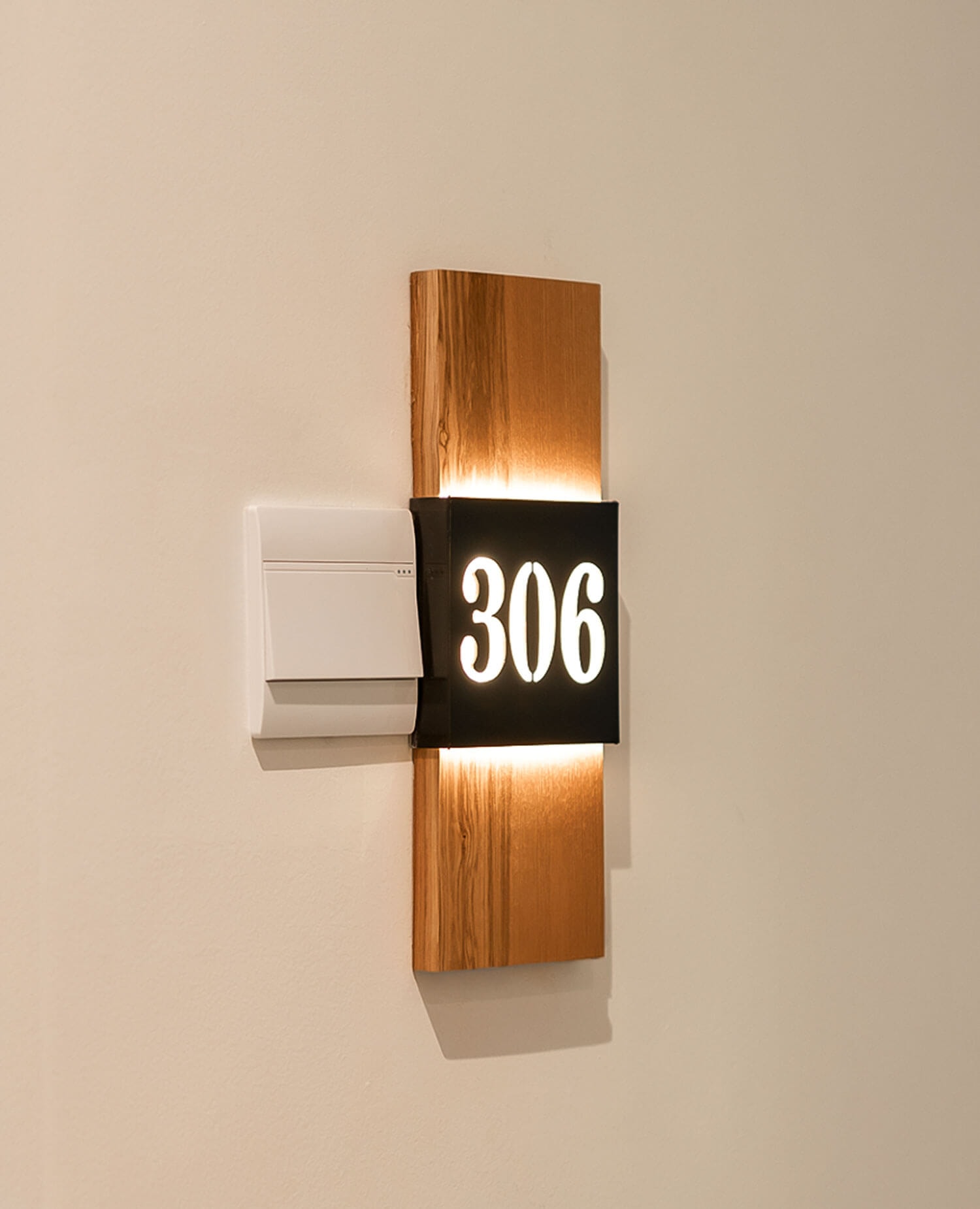 Nahaufnahme einer beleuchteten, an der Wand hängenden Zimmernummer '306' auf einem Holzuntergrund und mit Lichtschalter daneben.
