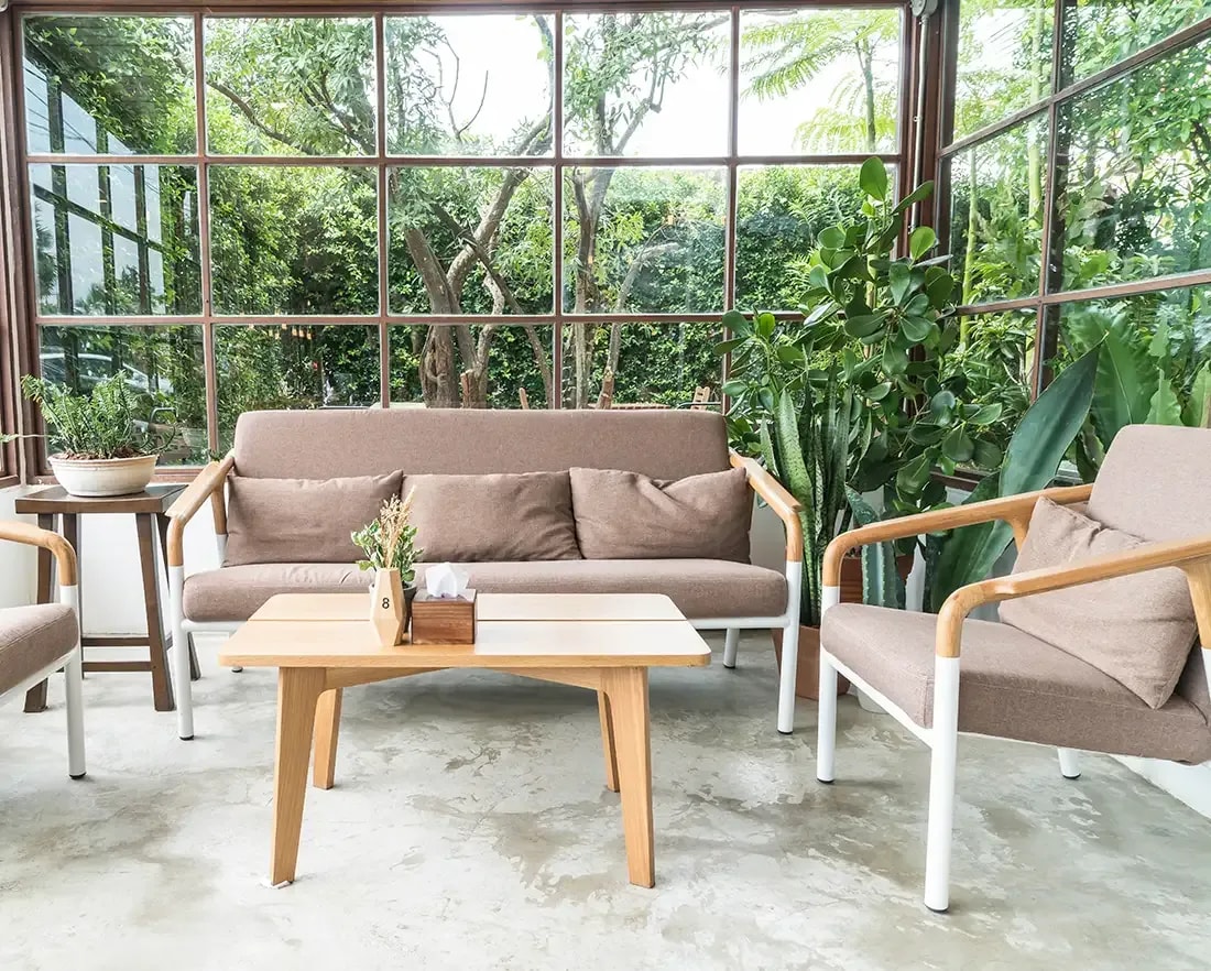 Photo of a conservatory with a light beige seating area in front of a glass front, through which lush garden greenery is visible