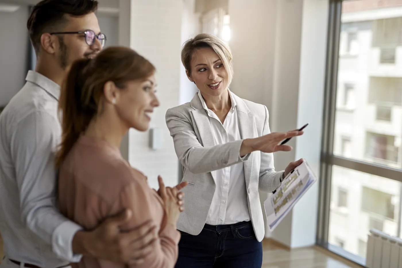 A female real estate agent in conversation with a an arm-in-arm couple, pointing out details of the apartment.