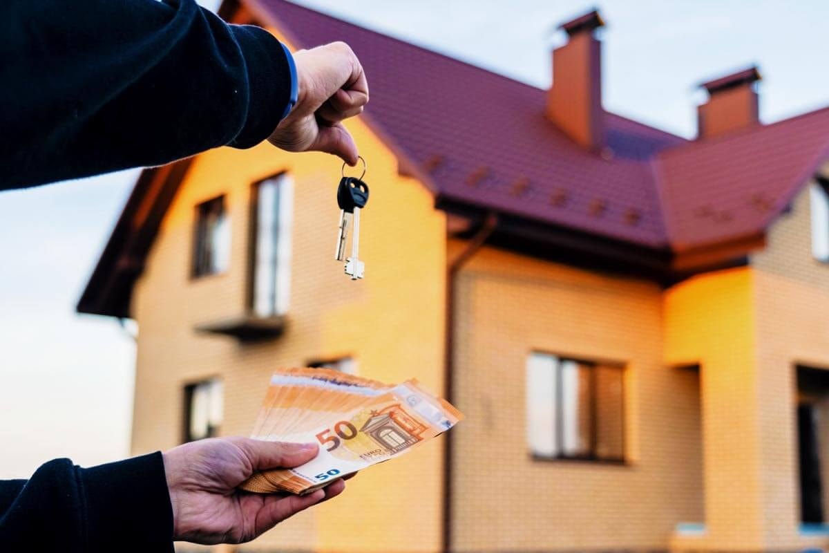 Hands holding a house key and banknotes in front of a residential building; symbolic image of a rental deposit and real estate transaction.