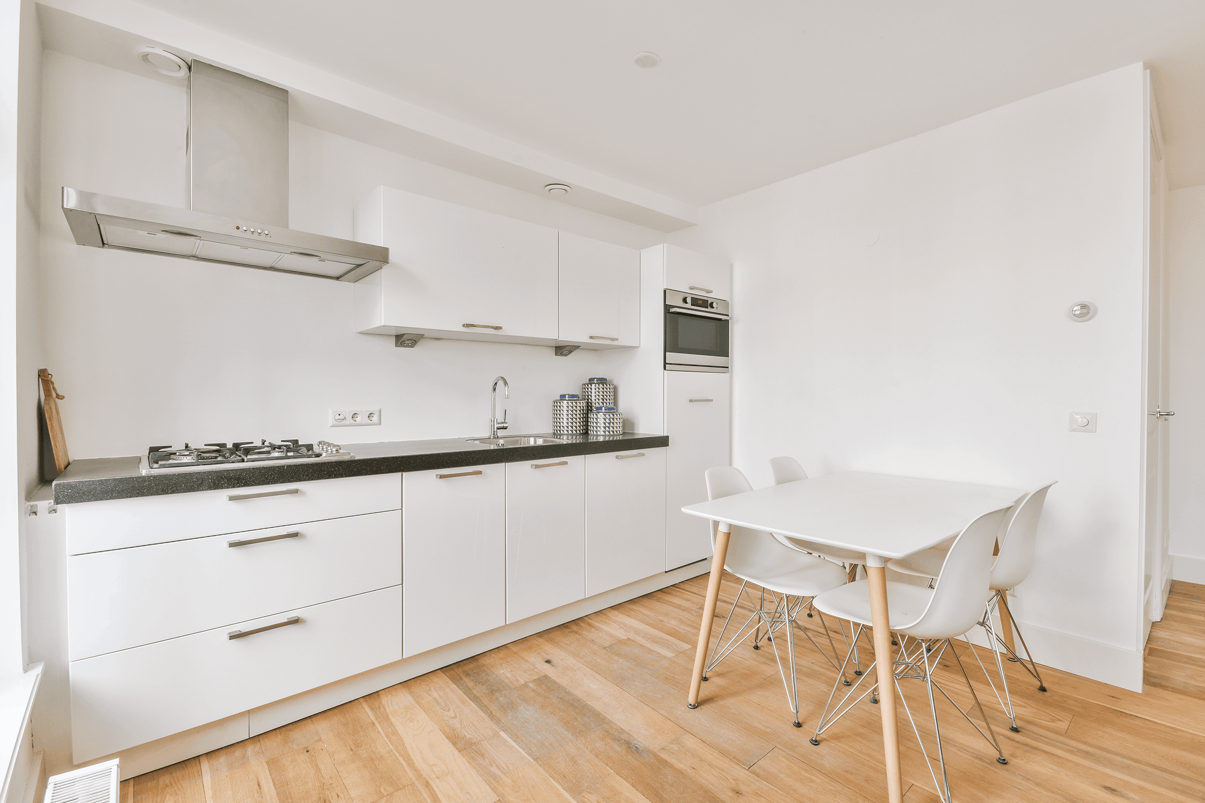 Simple white kitchen with dark worktop, gas hob and built-in oven, with a small white dining table and chairs alongside.