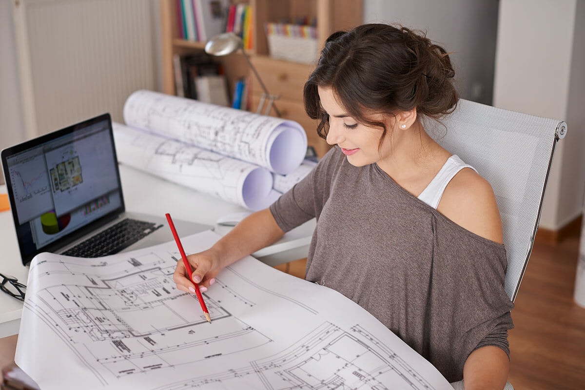 A graphic designer sits at a desk with a laptop and floor plans spread around, noting changes on an unrolled plan in front of her with a pencil.