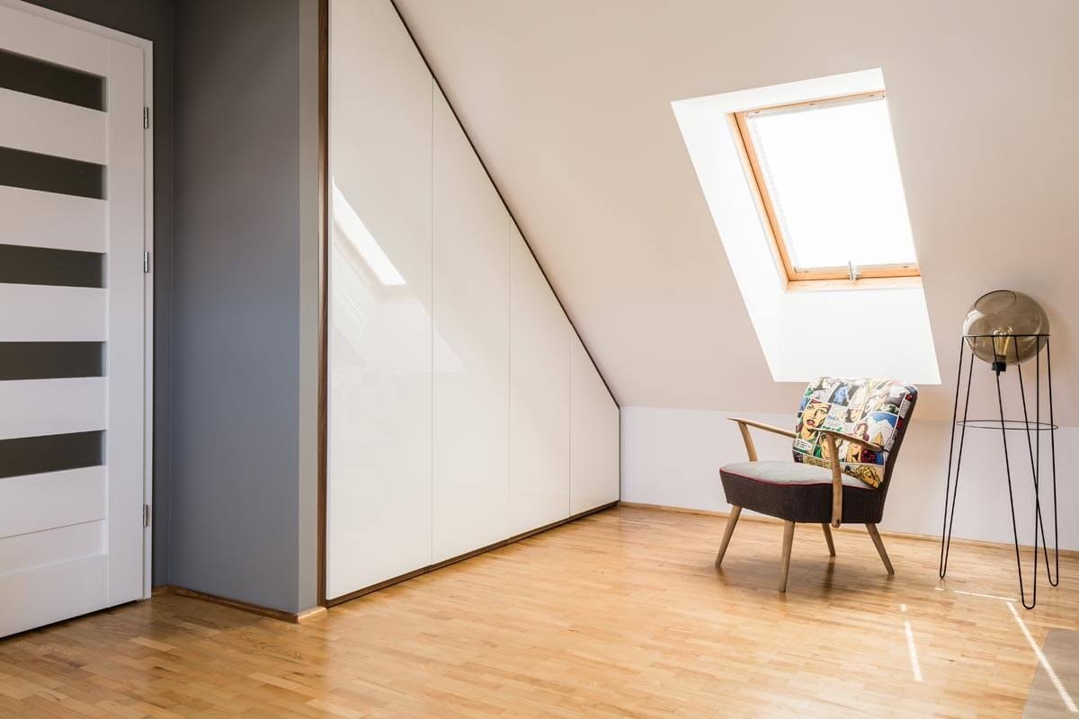 Blick auf die Ecke eines Raumes mit Dachschräge, in dem ein Sessel und eine Lampe unter einem Dachfenster stehen und links eine Zimmertür neben einem Einbauschrank ist.