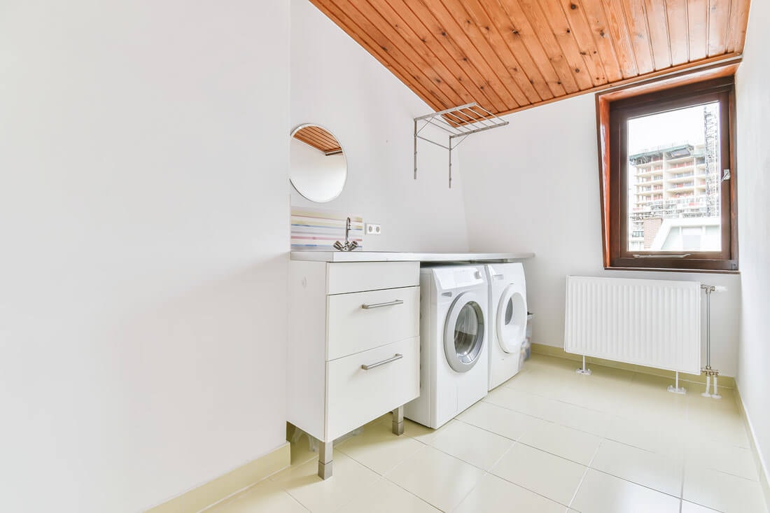 View from the entrance of a utility room toward a cabinet with a sink, a dryer and a washing machine, with a window on the opposite side through which the neighboring house is visible