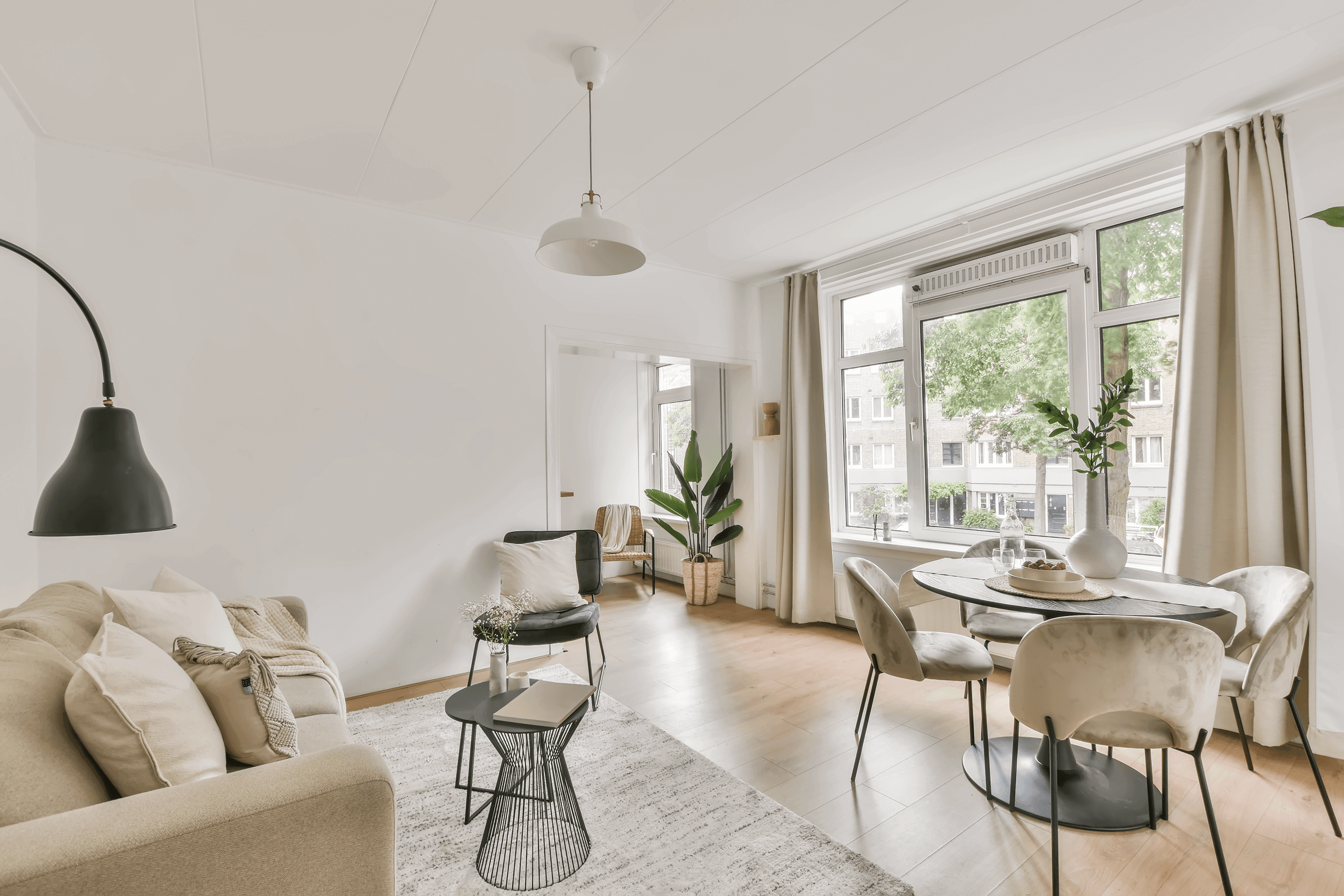 Bright, Scandinavian-style living room with a beige sofa, black pendant lamp, round dining table and large windows overlooking greenery.