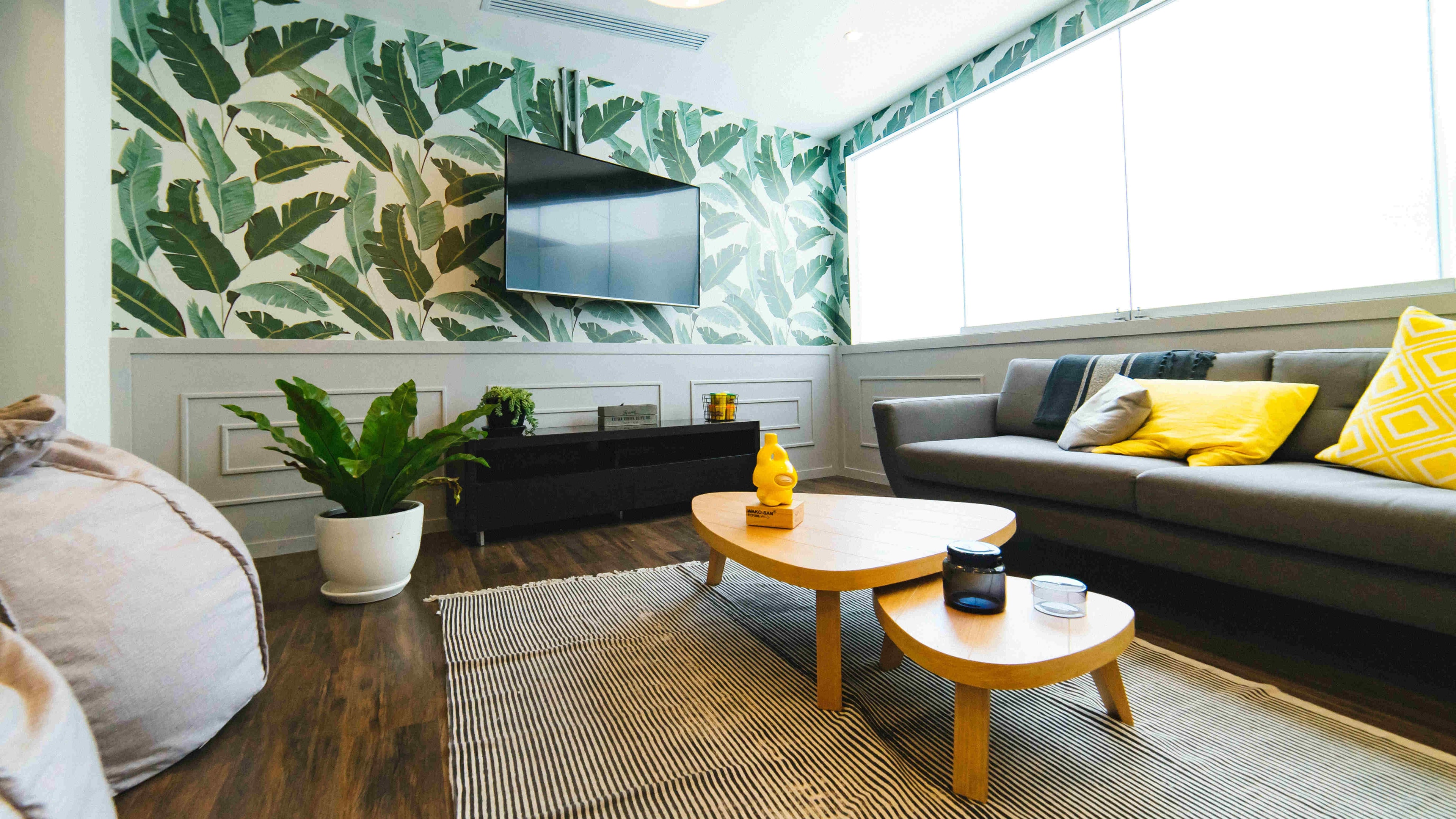 Corner view into a modern living room in earthy and green tones, with eye-catching leaf-patterned wallpaper and a very wide window front.