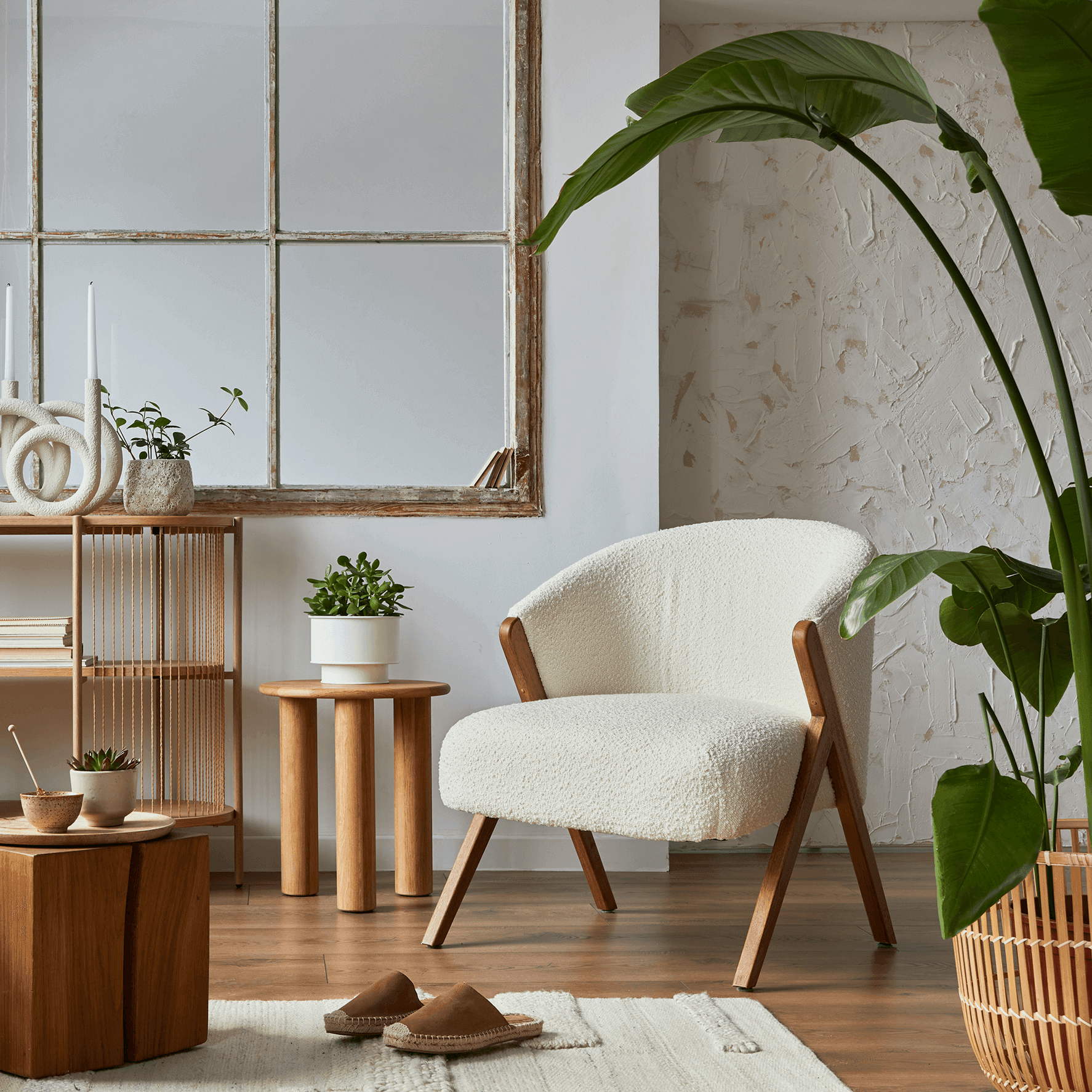 Living room corner in boho style with a white bouclé armchair, large indoor plant, wooden shelving unit with decorative objects and a vintage mirror.