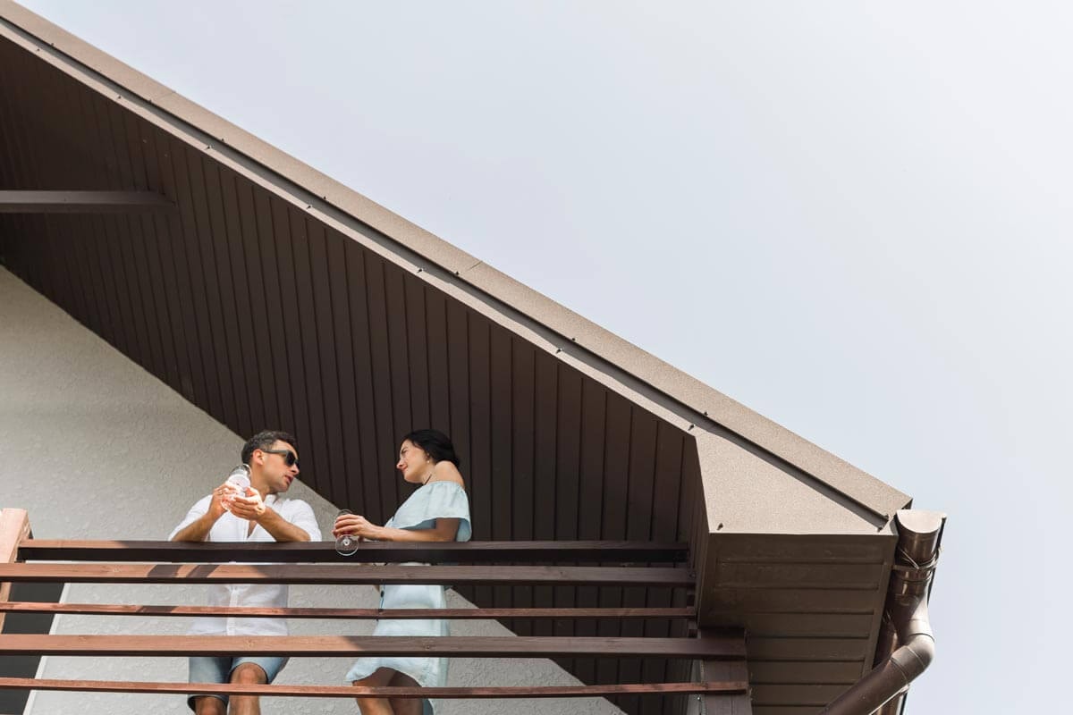 A couple stands on the balcony of an attic apartment beneath a dark roof overhang, chatting; the man in a white shirt with sunglasses, the woman in a light blue top — shot from a low angle.