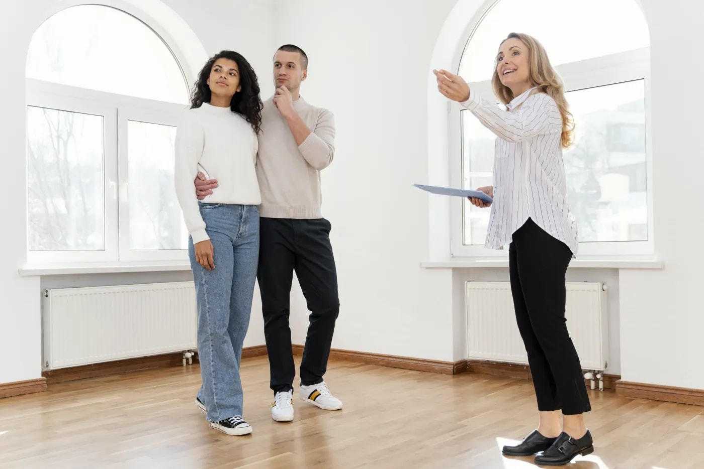 A female real estate agent stands with a couple in an empty apartment, pointing to a detail behind the viewer.