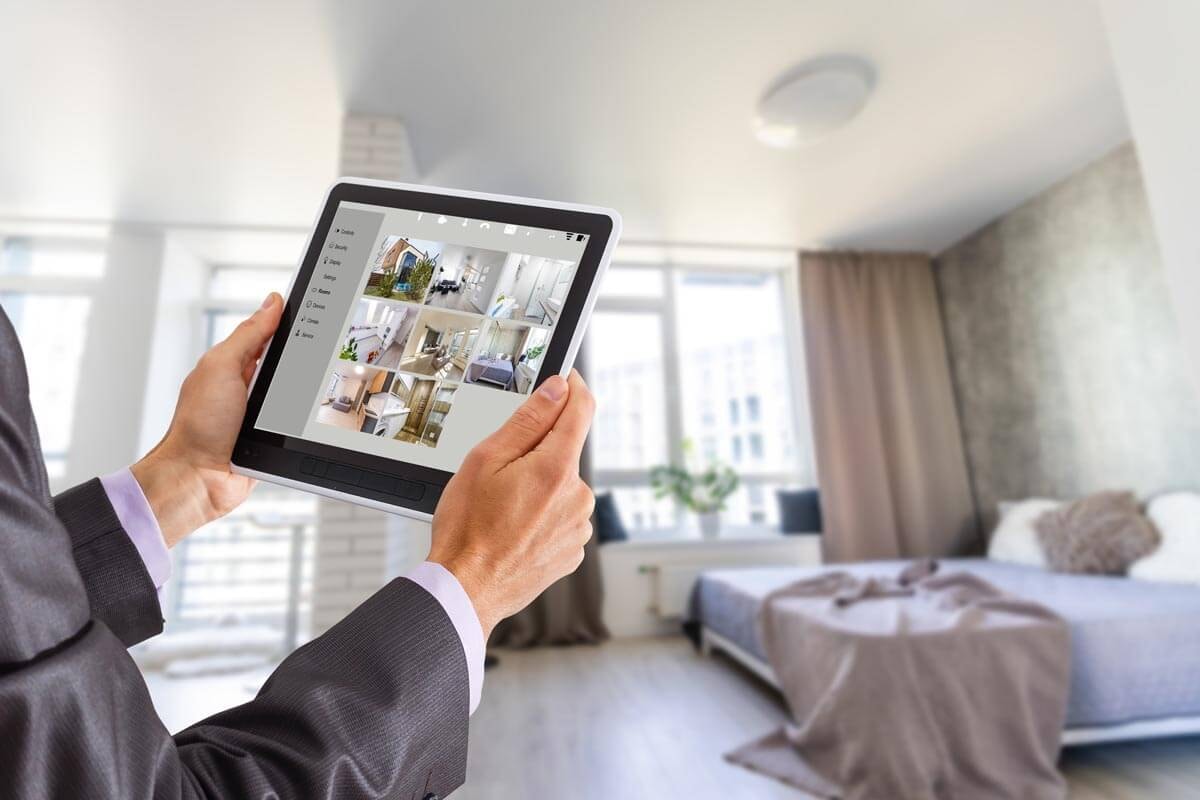 A man holds up a tablet with both hands showing an overview of photos of an apartment, while a bedroom is visible in the background and neighboring high-rise buildings can be seen through the windows