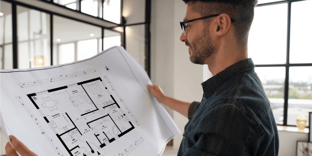 An architect in their studio looks contentedly at a dimensioned floor plan they are holding up with both hands