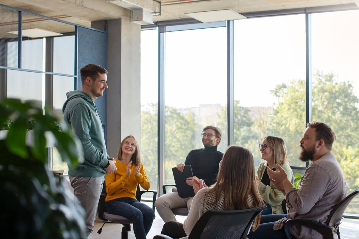 Various people seated in a semicircle of chairs in front of floor-to-ceiling windows showing trees outside; one person is standing and speaking to the others.