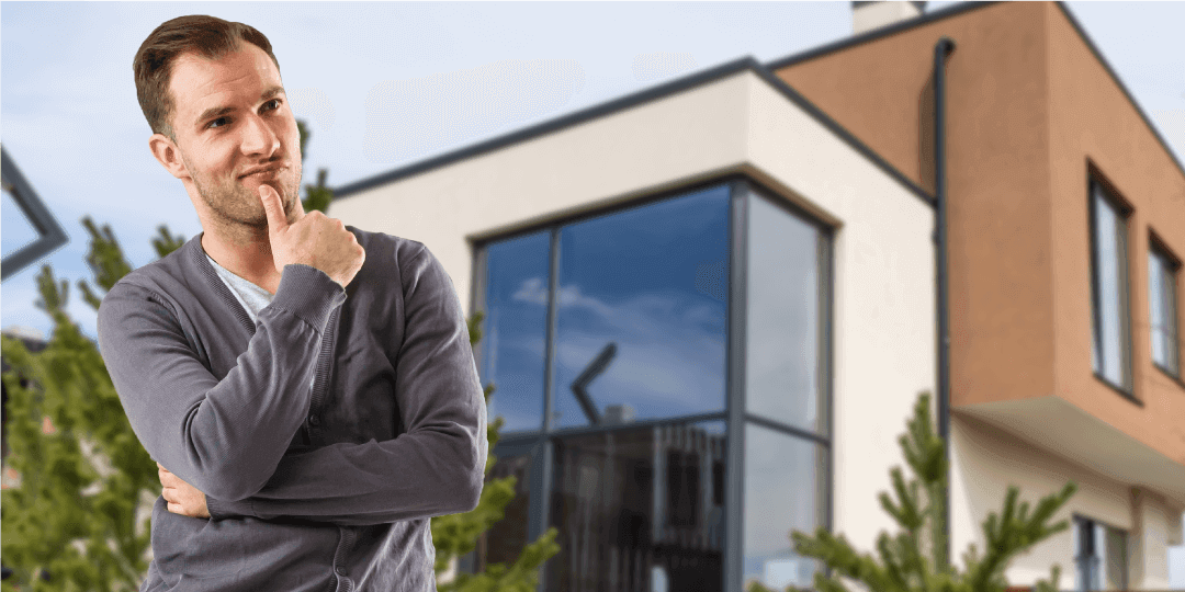 A man stands with a thoughtful expression, thumb resting against his chin, in front of a modern flat-roofed building