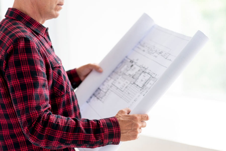 A man in a red-and-black checked shirt holds a construction drawing in front of him with both hands