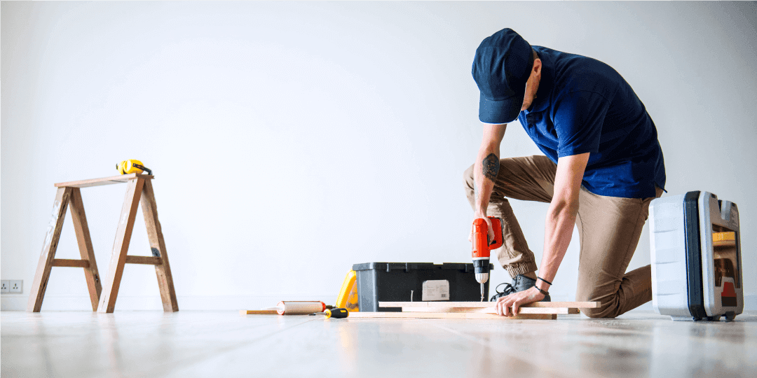 A tradesman works on floor panels in an empty room with a drill, its case lying next to him, and a wooden sawhorse with a tape measure on it standing a step further away