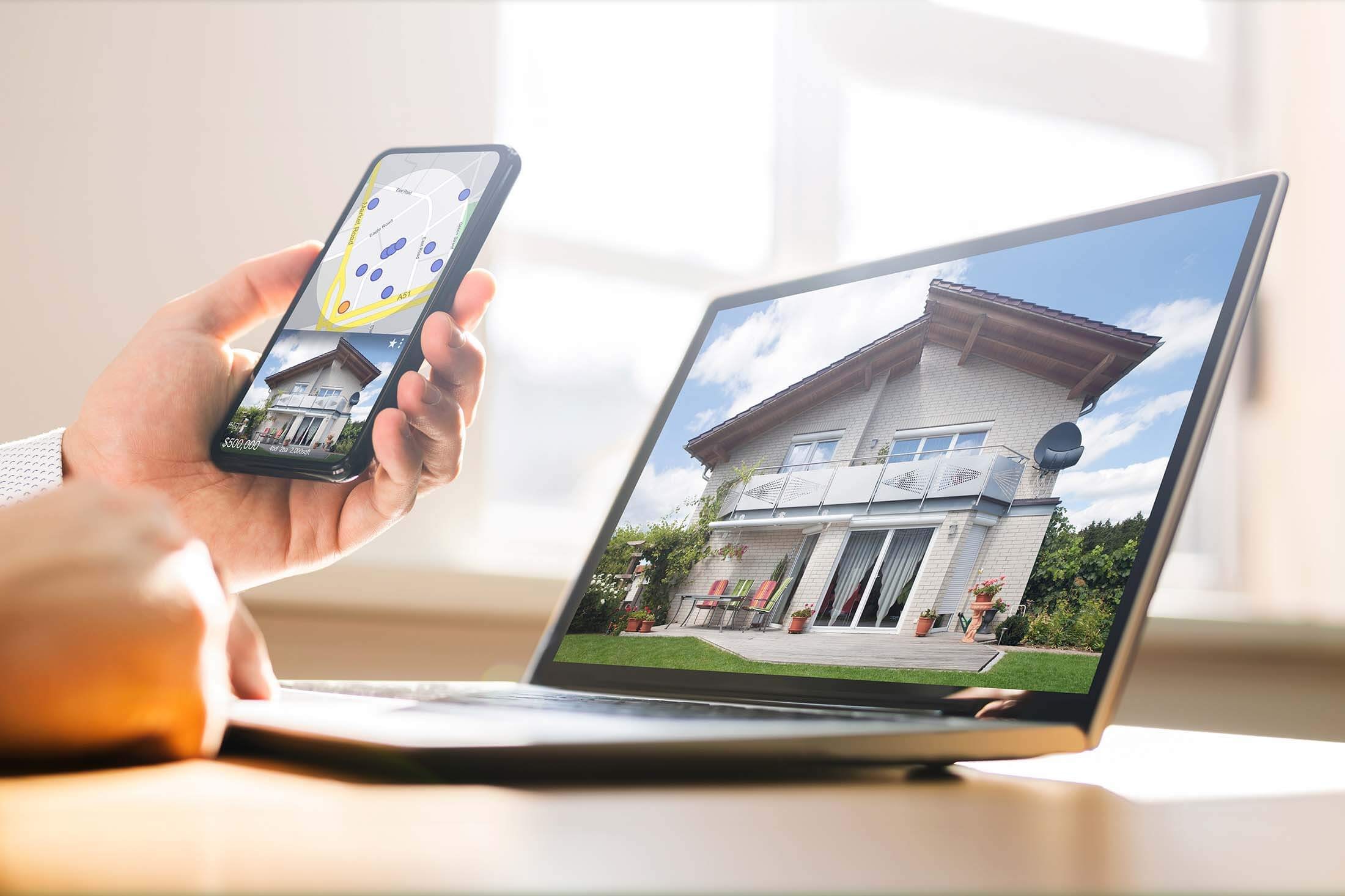 A person sits at a table comparing property data in a phone app with the image on an open laptop standing on the table in front of them