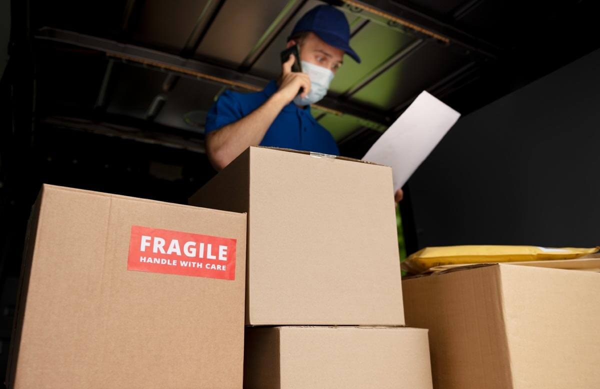 A delivery worker in blue workwear and a face mask stands in the dark loading area of a van, talking on the phone while holding a document; in the foreground a box with a red Fragile sticker.