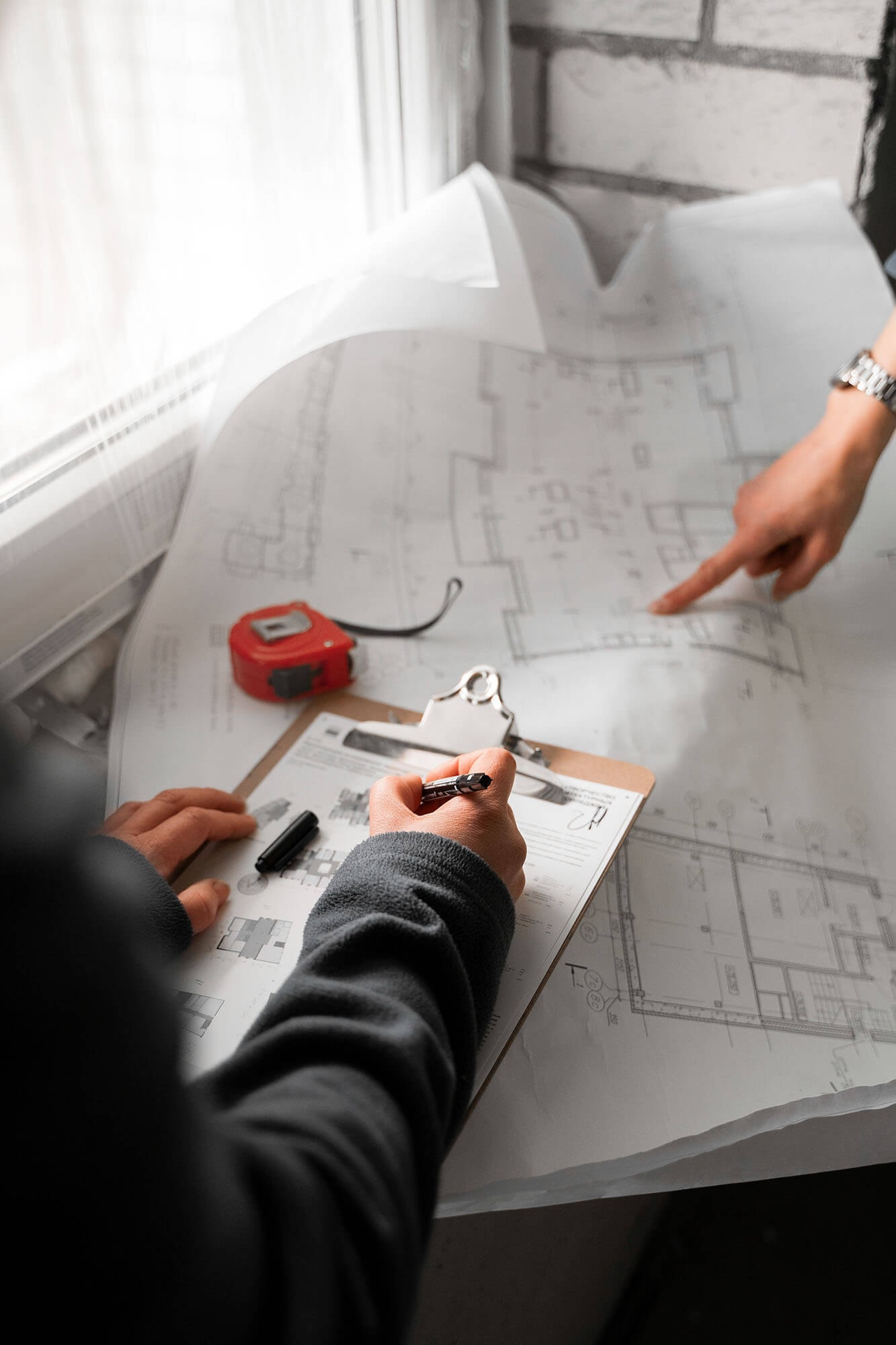A floor plan lies open and crumpled on the windowsill of a building shell, with a hand pointing to a detail while a clipboard and measuring tape rest beside it.