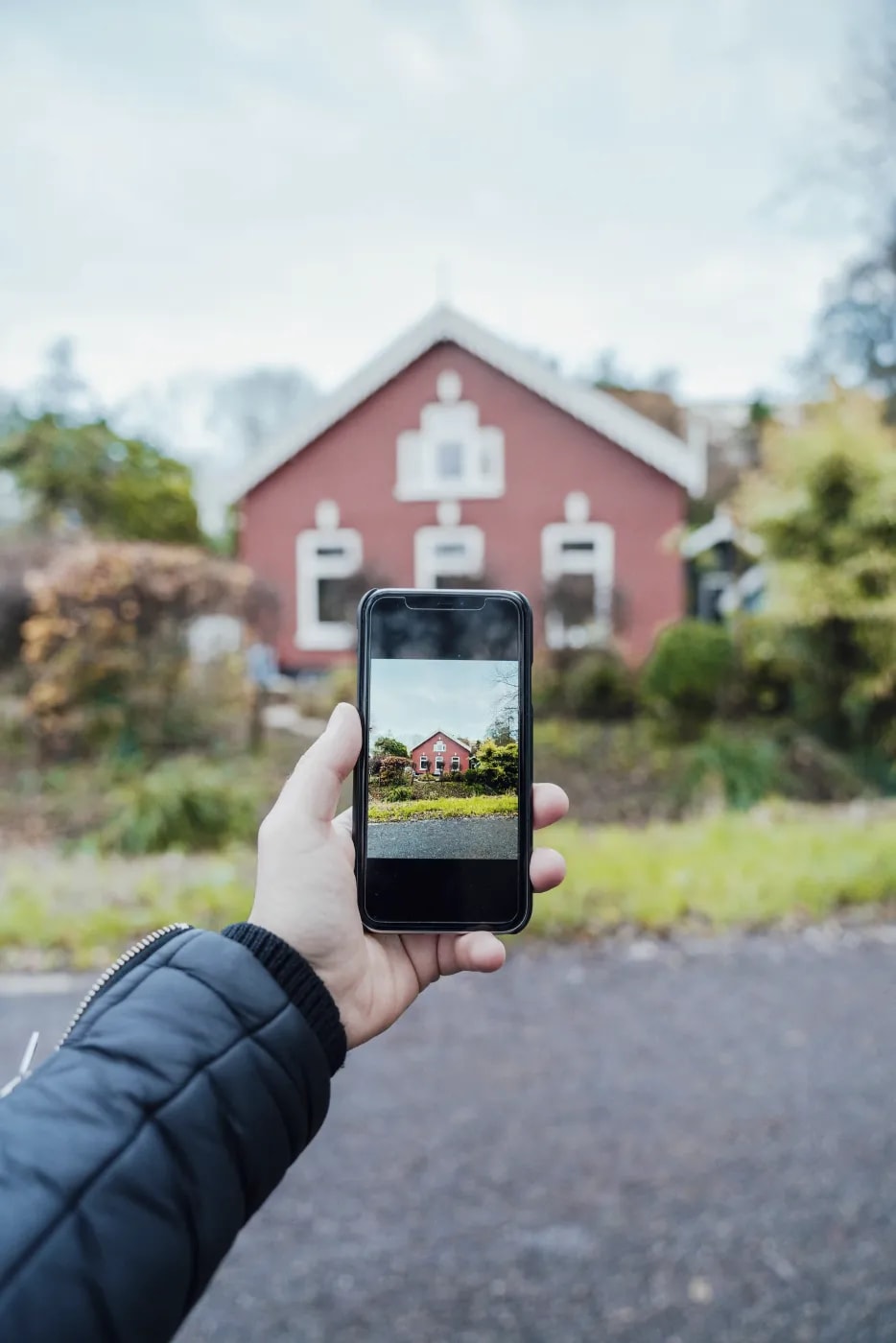 Close-up of a hand holding a phone taking a photo of a small single-family house with a reddish facade and white window and door frames