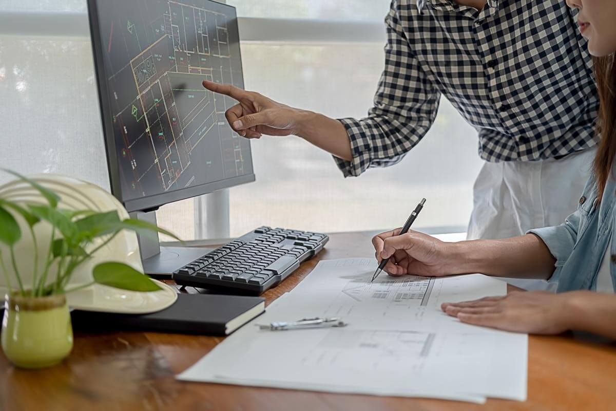 One hand points to something on a blueprint on a monitor, while a second hand takes notes on a facade drawing lying in front of the monitor