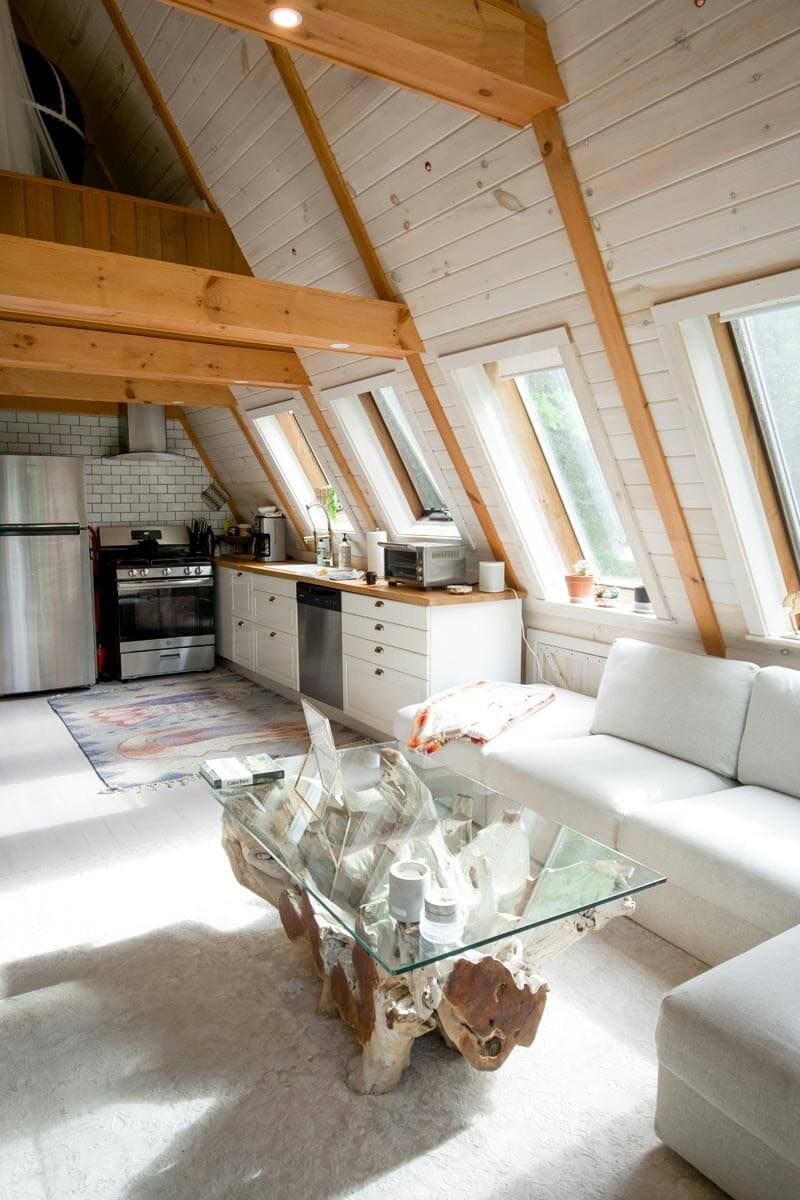 A converted attic with striking A-frame natural timber beams and white wall planks, inside an open kitchen with white base units and stainless steel appliances on the left, a white seating area and a coffee table with a tree-root base and glass top on the right.