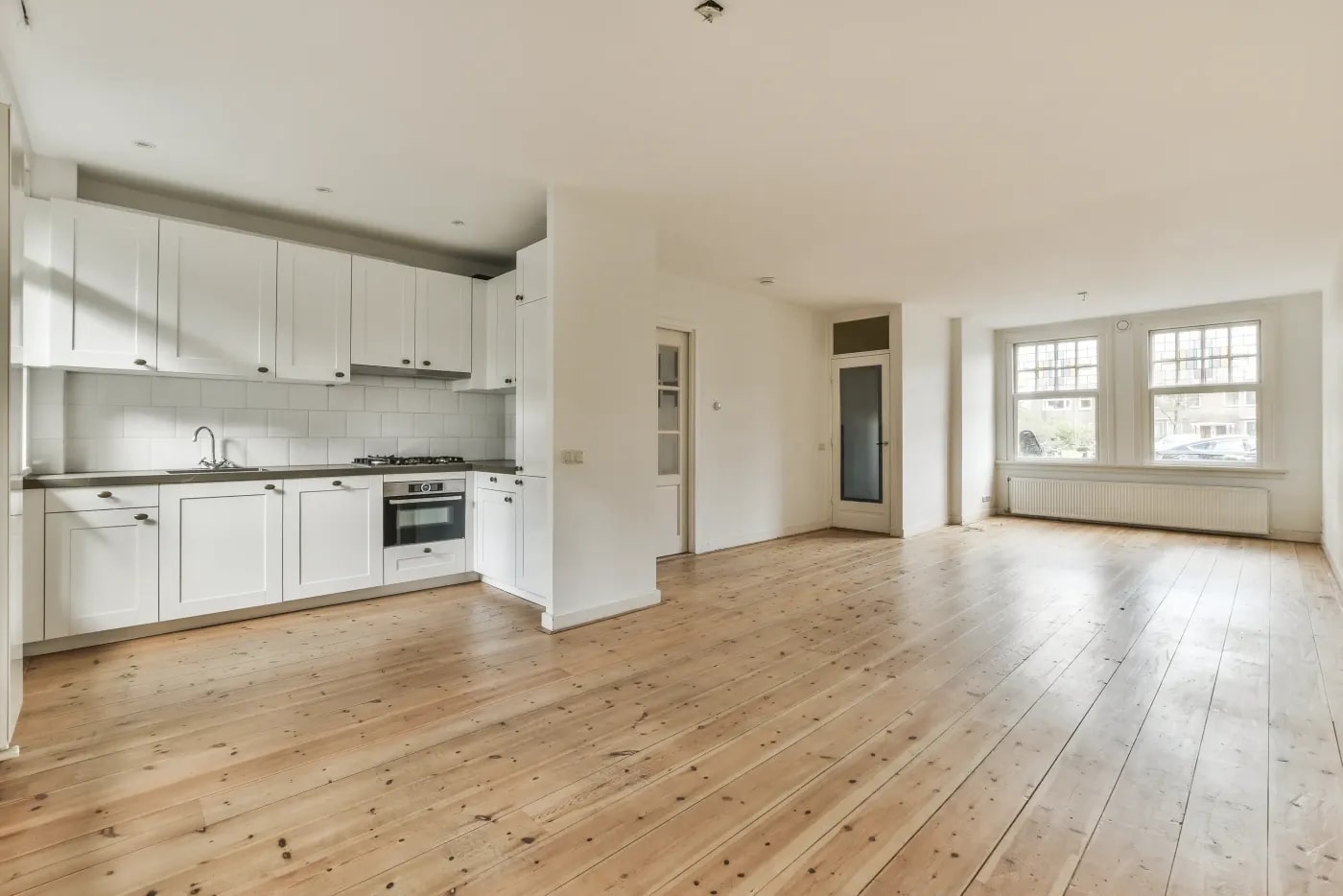 A very large living room with wooden flooring, an open kitchen counter and windows on each of the short sides.