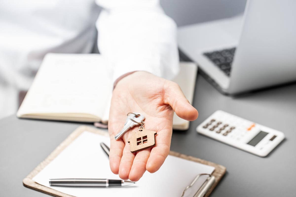 Close-up of a hand offering a house key with a keyring in the shape of a small house; beneath it some documents and a calculator.