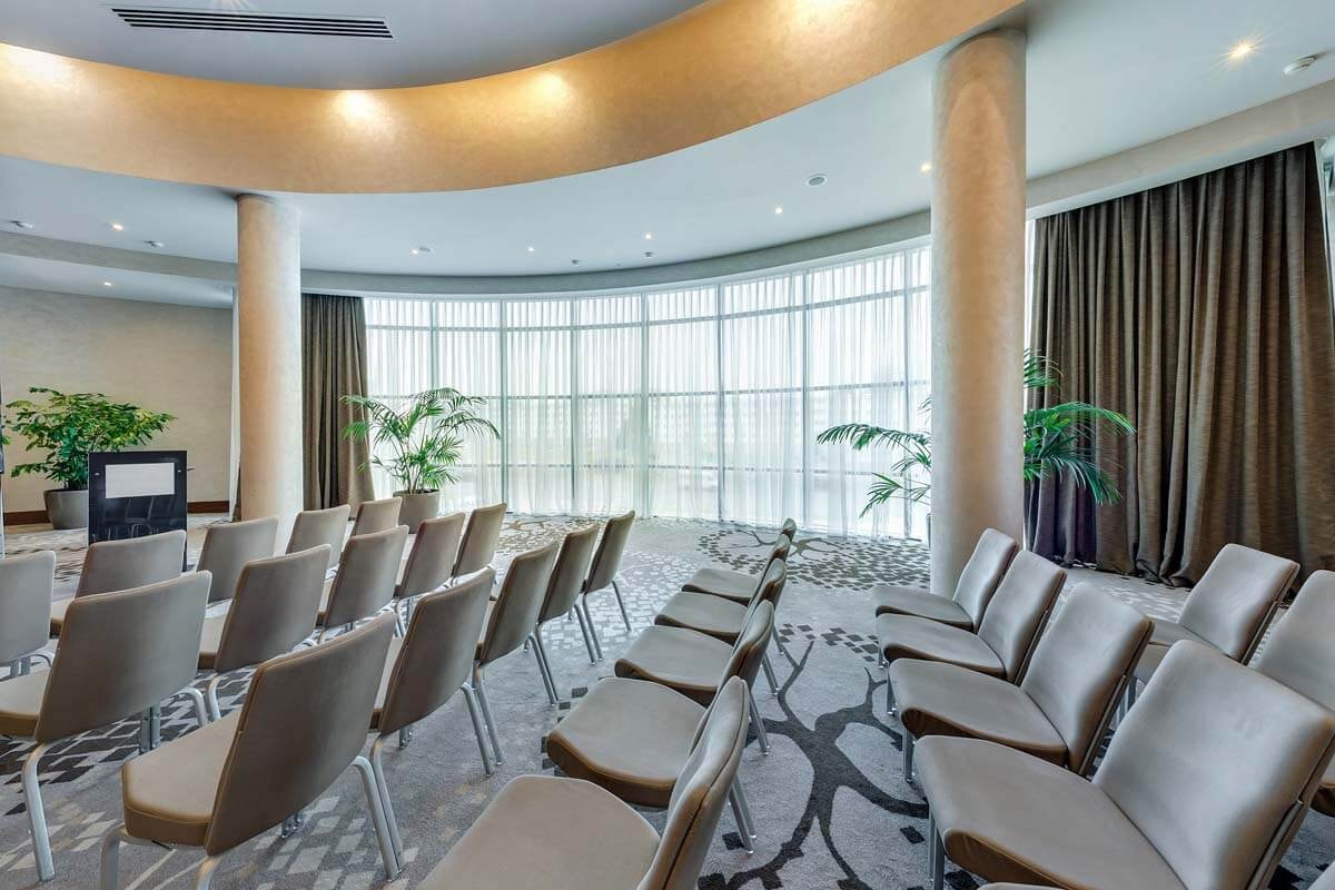 Side view of rows of chairs in a brightly lit lecture hall with two columns and a wide window front with net curtains and drapes in the background.