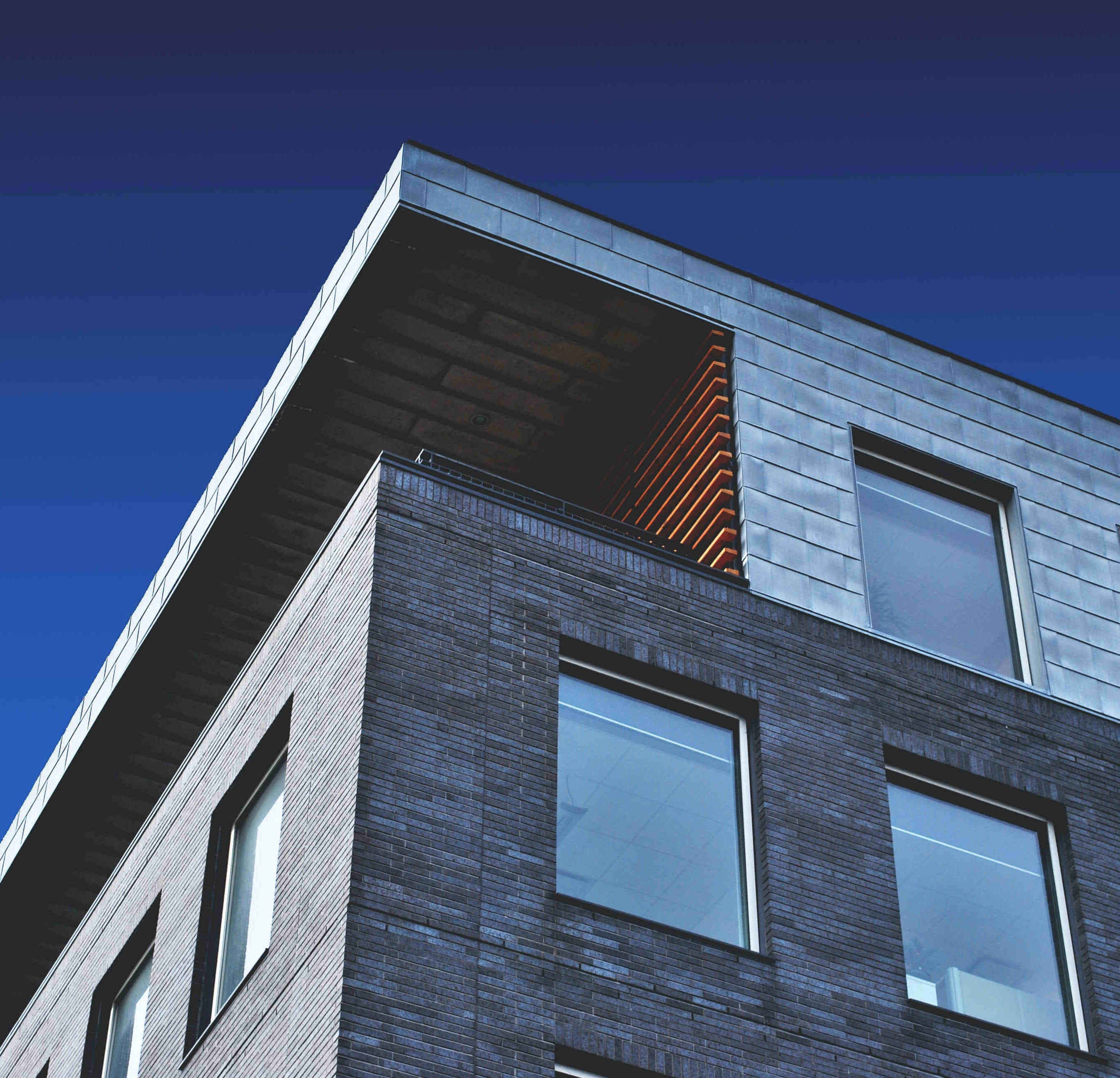 View from below of the upper corner of a house with a flat roof, balcony and many windows on the facade.