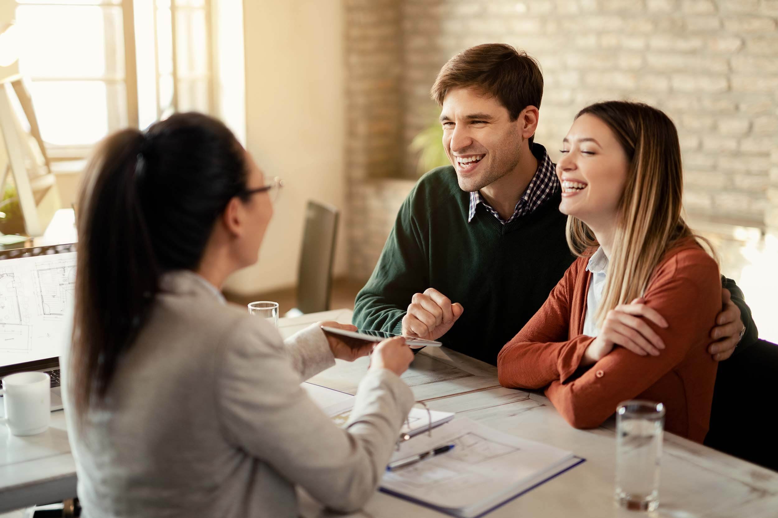 A real estate agent sits across from a laughing couple at a desk with building documents on it, showing them something on a tablet