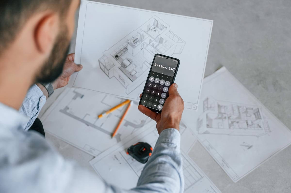 Rear view of a man holding a calculator over 3D floor plans on a table, looking at the display showing 29620 + 1540