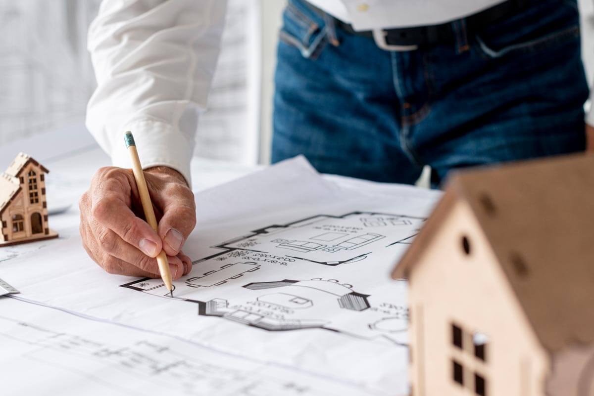 Close-up of a person standing at a table with a large floor plan alongside two wooden miniature houses, making notes on it with a pencil.