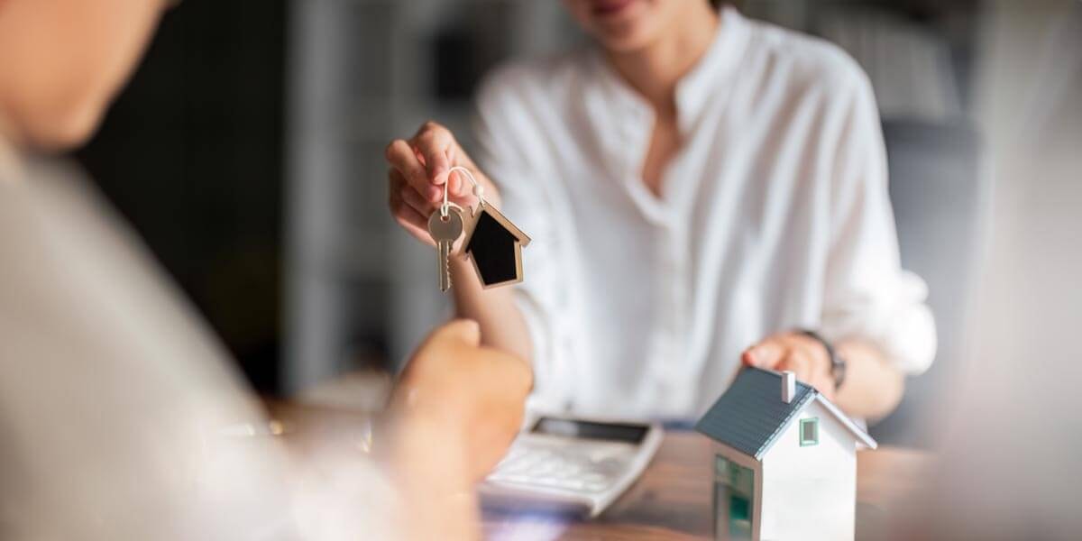 A female estate agent sits at a table with a client and hands over a front door key; next to them a miniature house with a light blue gabled roof.