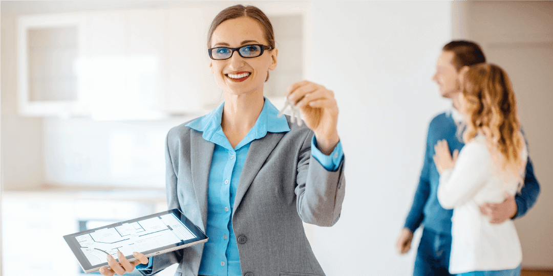In the foreground, a real estate agent is shown holding a tablet with a floor plan in one hand and holding up an apartment key with the other, while in the background an arm-in-arm couple contentedly look at the open kitchen they are standing halfway inside