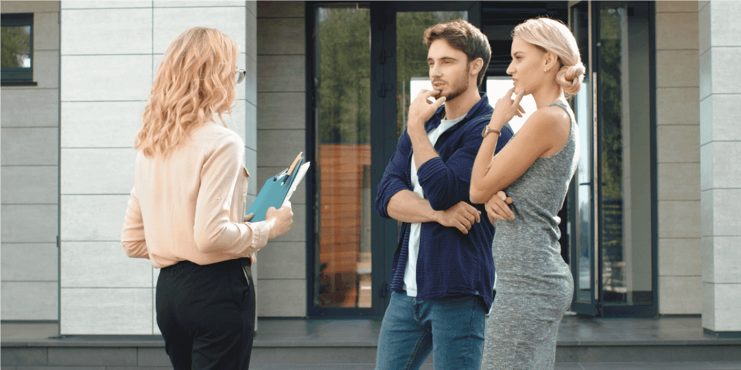 A real estate agent stands with two prospective buyers in front of a house entrance, explaining something to them while they listen attentively and thoughtfully