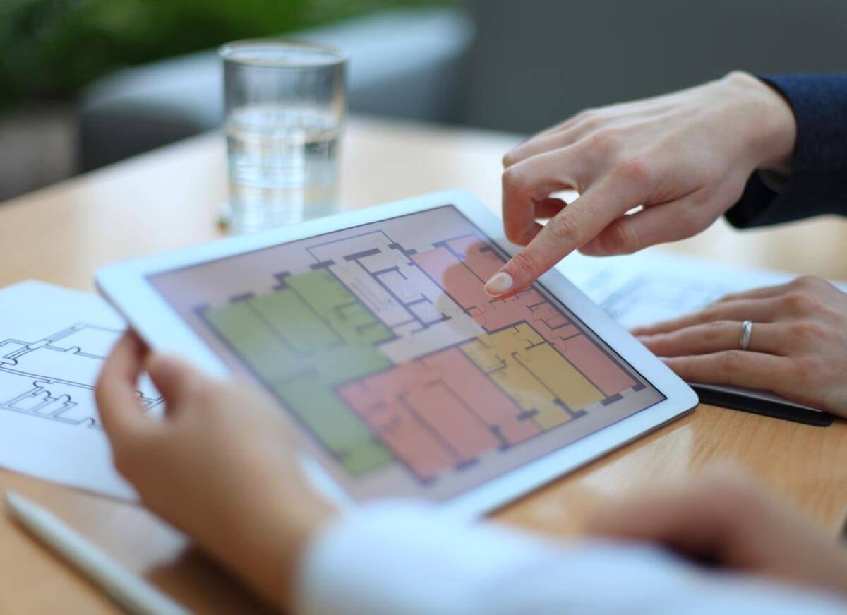 One hand holds a tablet showing the floor plan of a multi-family building with a stairwell, in which the individual residential units are colour-coded, while a second hand points with the index finger to a detail of the floor plan in one of the units.