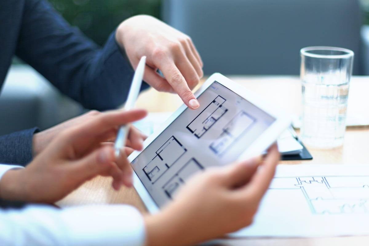 Hands at a desk hold a tablet and a pen, while a third hand points to one of four floor plans shown on the tablet